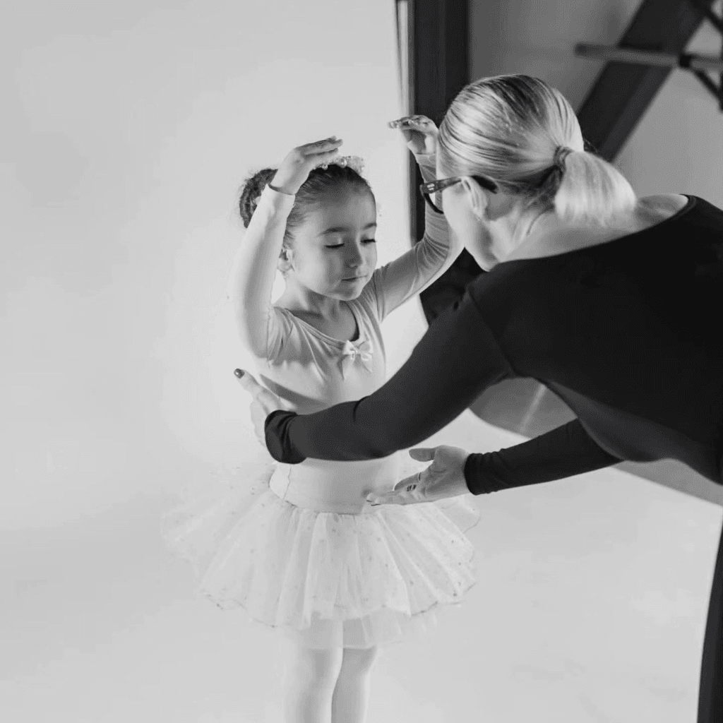 A ballet teacher gently adjusting a young ballerina's arms and posture in the studio